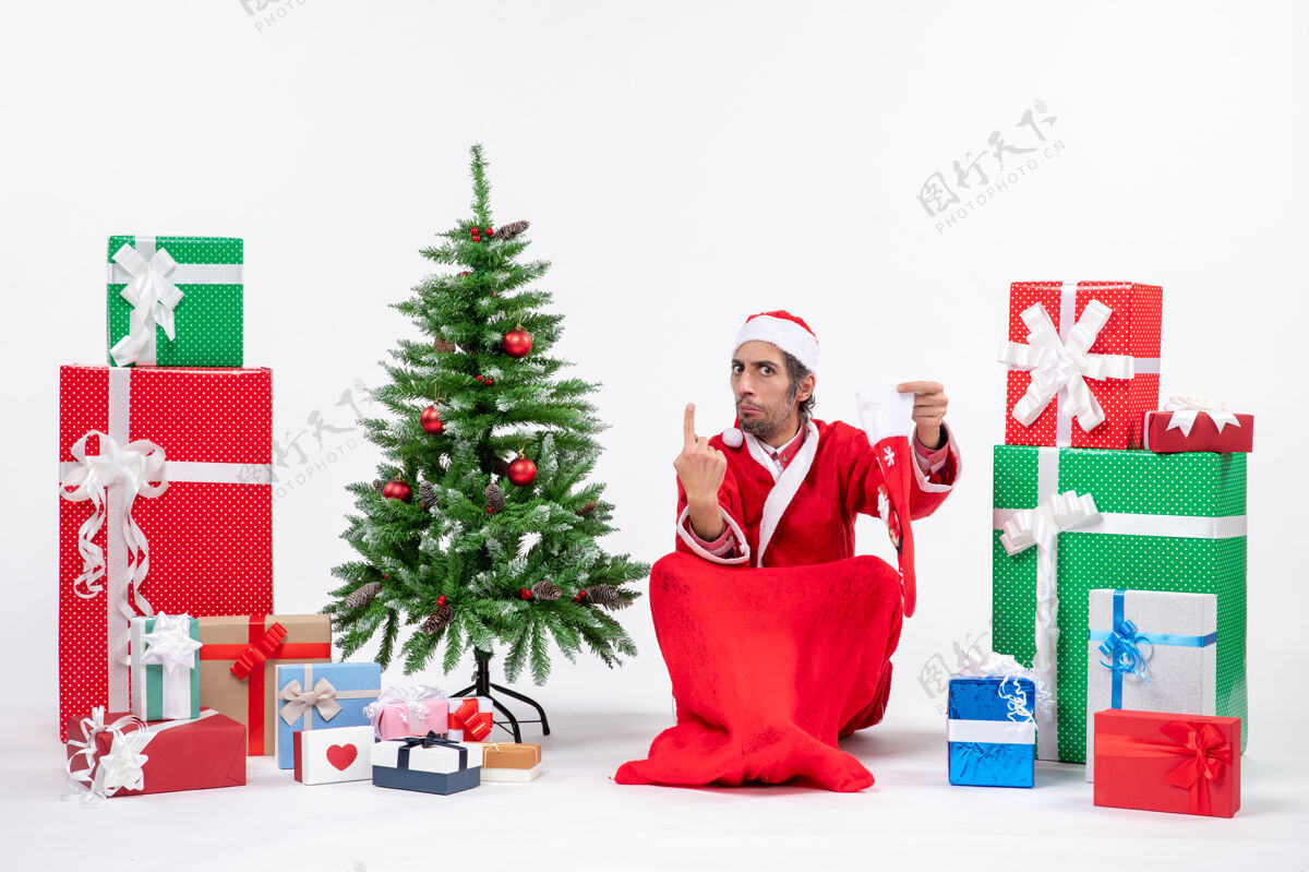 坐着紧张的圣诞老人展示了一个坐在地上 拿着圣诞袜子靠近礼物和装饰在白色背景上的新年树礼物圣诞老人礼物