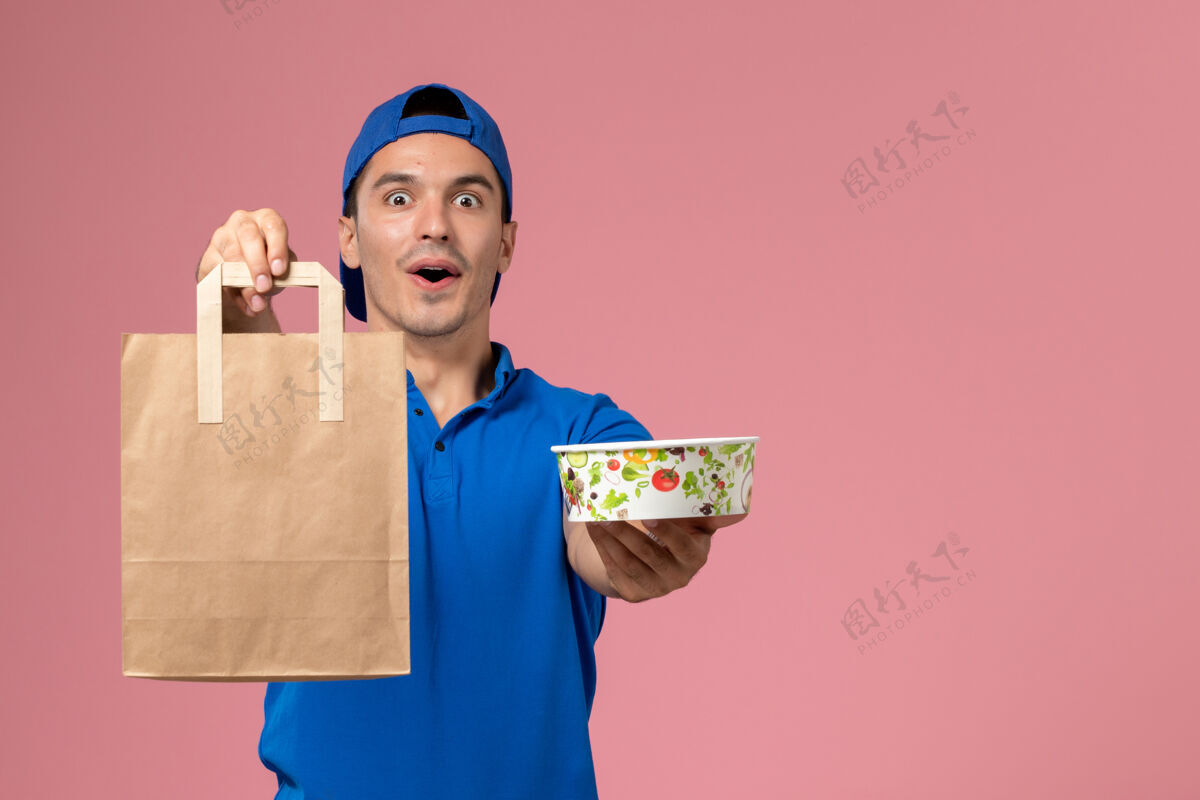 成人正面图：身穿蓝色制服和斗篷的年轻男性快递员 手上拿着快递包裹和碗 放在粉红色的墙上年轻粉色碗
