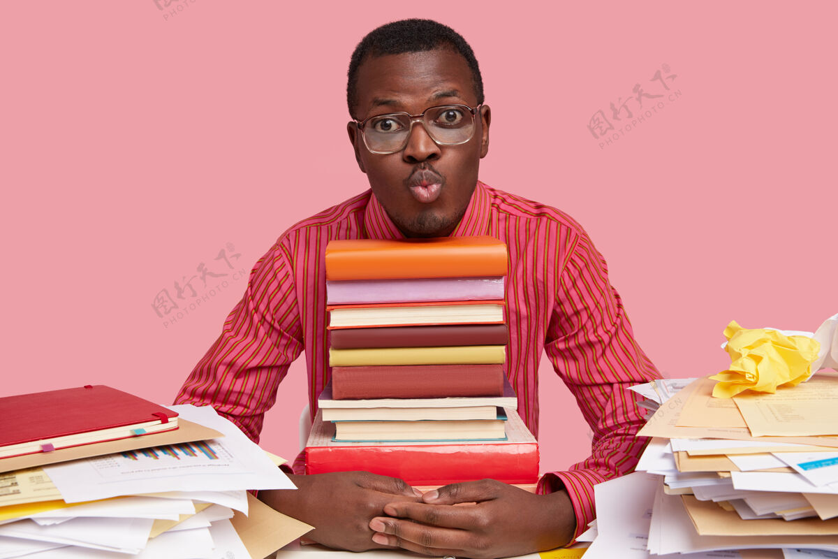 书籍英俊的黑皮肤男人撅着嘴 拿着一堆书 桌面上乱七八糟的 戴着眼镜和条纹衬衫眼镜教育独自
