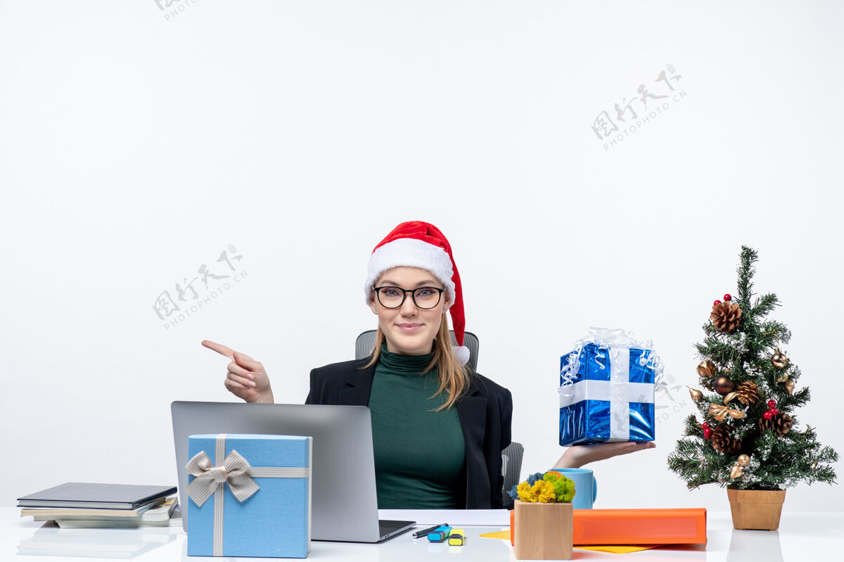 桌子带着好奇的惊喜微笑的年轻女子戴着圣诞老人的帽子戴着眼镜坐在桌子旁展示圣诞礼物指着右边的东西礼物年轻人一边