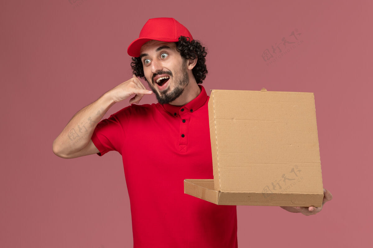 男性信使正面图：身穿红色衬衫和披风的男性快递员手持浅粉色墙上的送货食品箱服务披萨视图