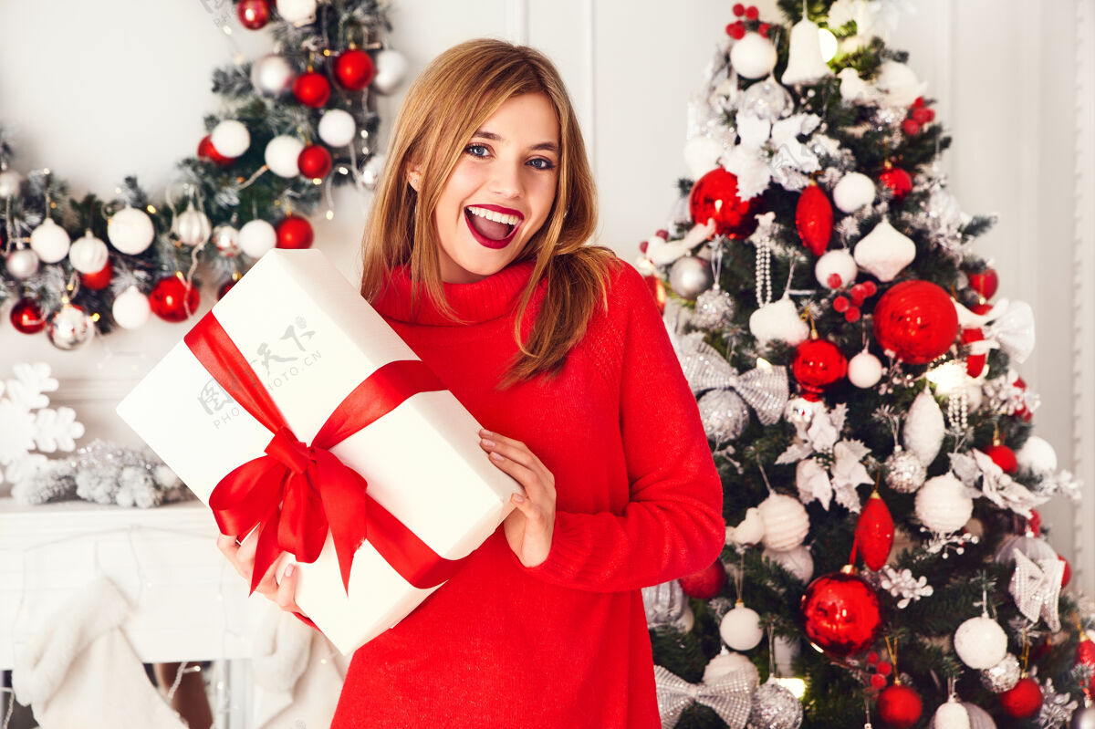 装饰微笑的女人拿着许多礼品盒在装饰好的圣诞树旁摆姿势毛衣惊喜青少年