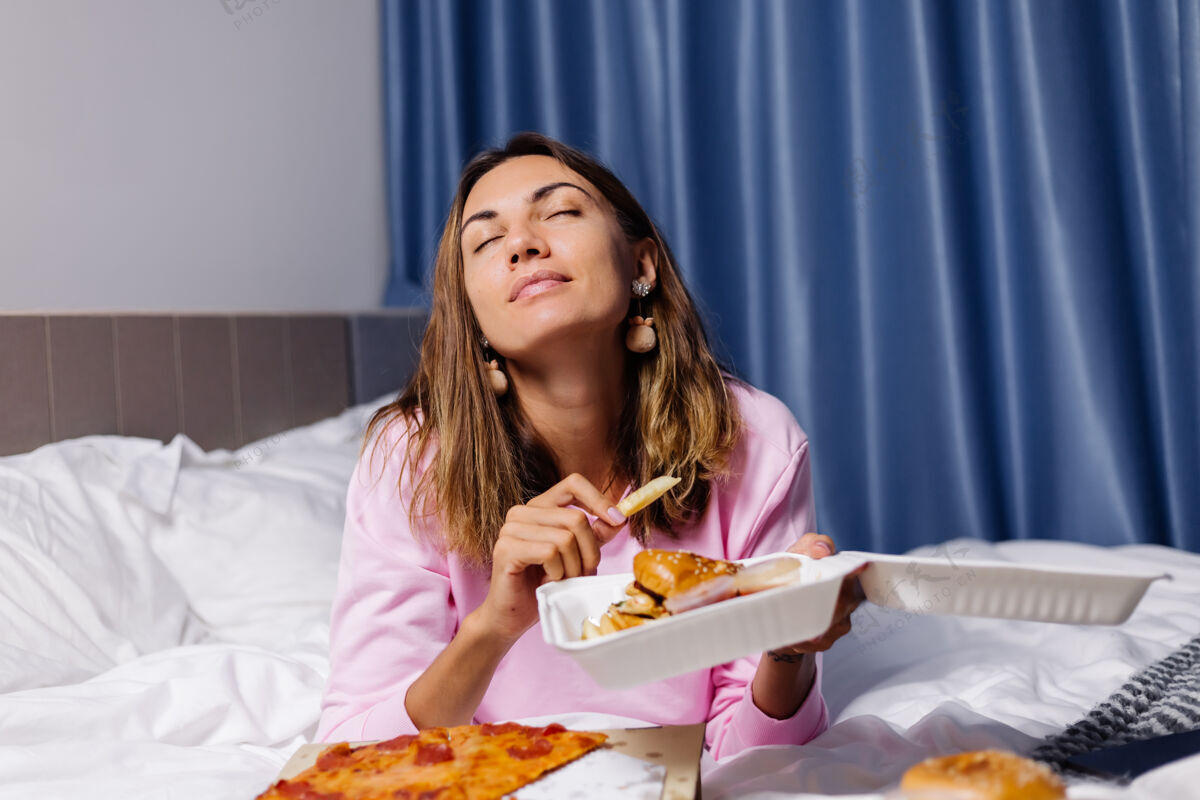 食物女人东快餐从床上交货在卧室在家里的女性享受脂肪食品比萨饼和汉堡渴望碳水化合物饿睡衣快餐