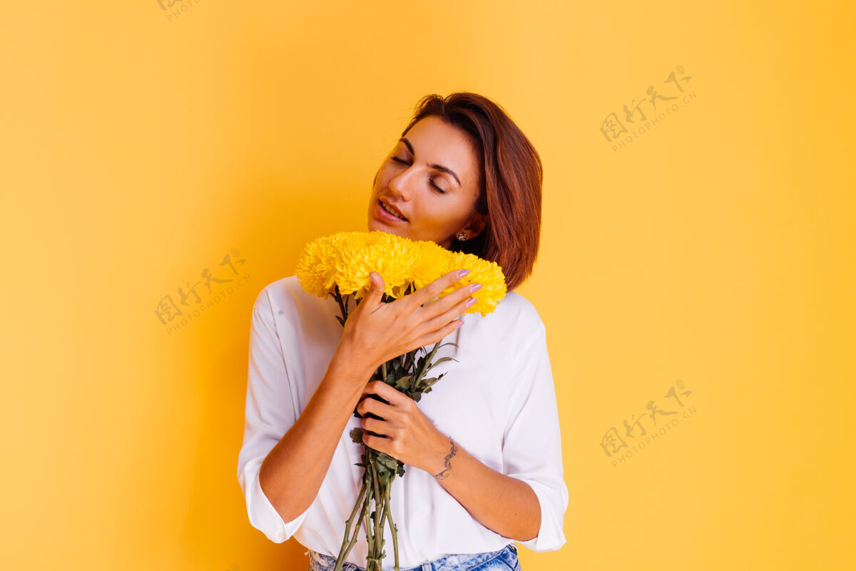 惊喜摄影棚拍摄的黄色背景快乐的白人妇女短发穿着休闲服白衬衫和牛仔裤手持一束黄色紫苑礼物期待年轻