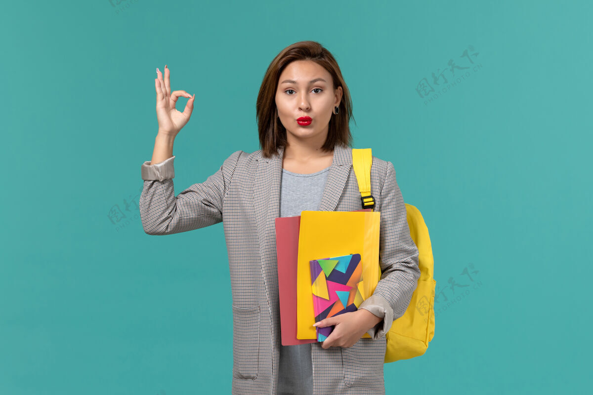 穿着身着灰色夹克 背着黄色背包 在蓝色墙上拿着文件和文案的女学生的正视图Female女学生正面