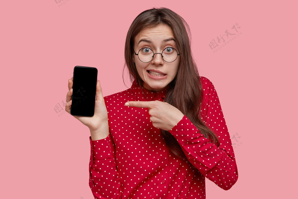 显示戴着眼镜的黑发少女困惑不解 指着模拟屏幕的电子产品 穿着红衬衫 宣传新设备 有一双绿色的眼睛电话青少年食指