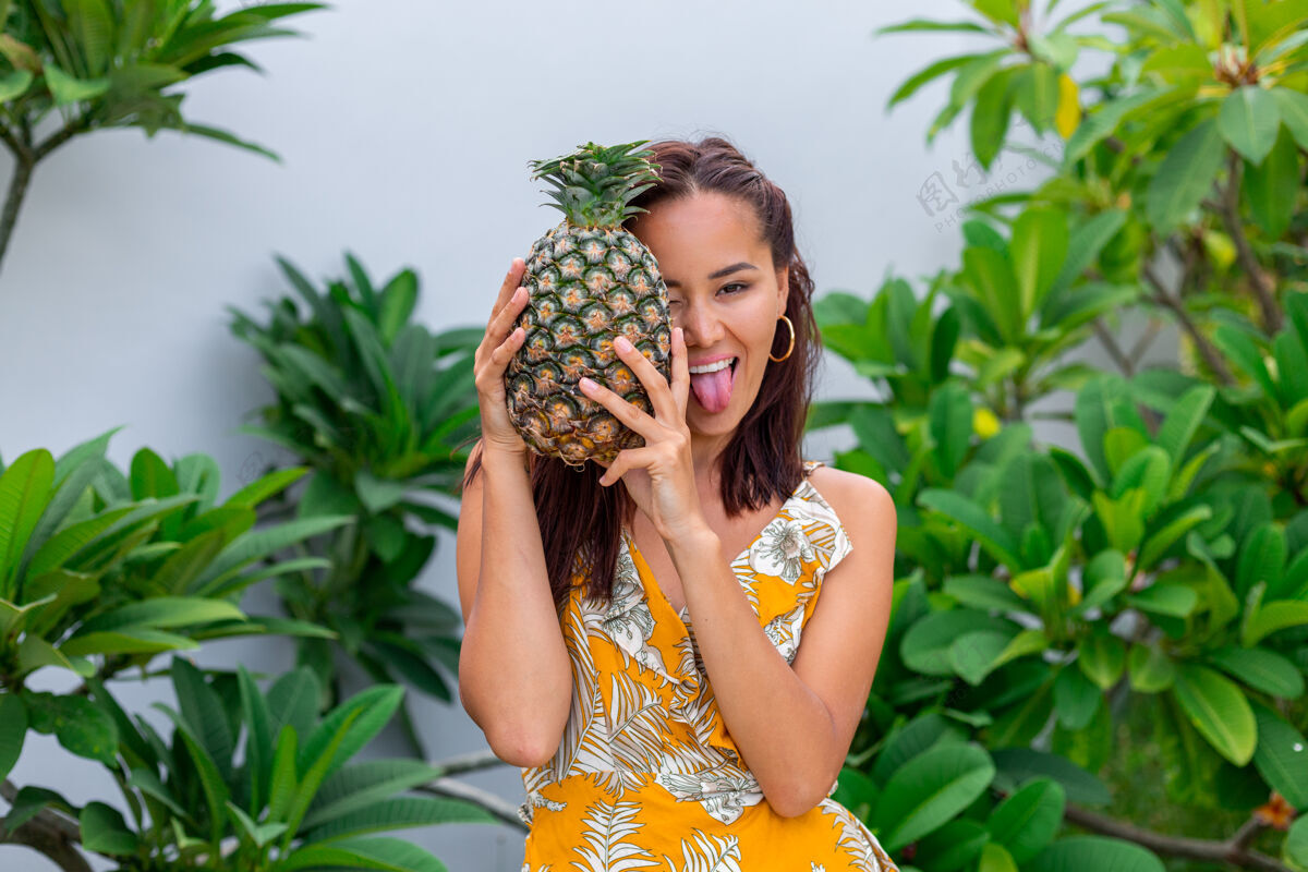 户外身着黄色夏装 手持菠萝的快乐亚洲微笑女人的画像舌头出来自然饮食