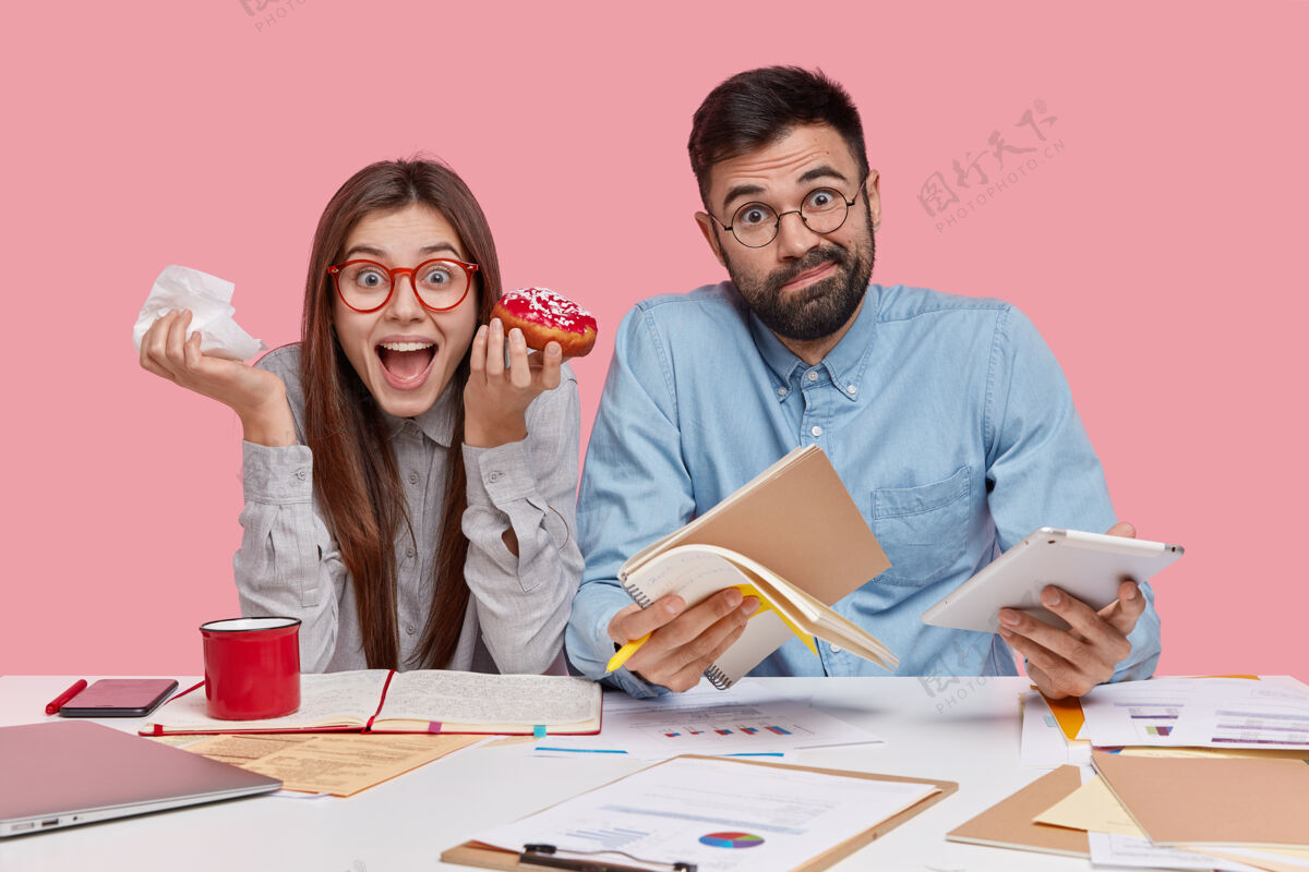 文件室内拍摄的男女同事吃着美味的甜甜圈 在笔记本上记下笔记 运用现代科技同事笔记本电脑惊讶