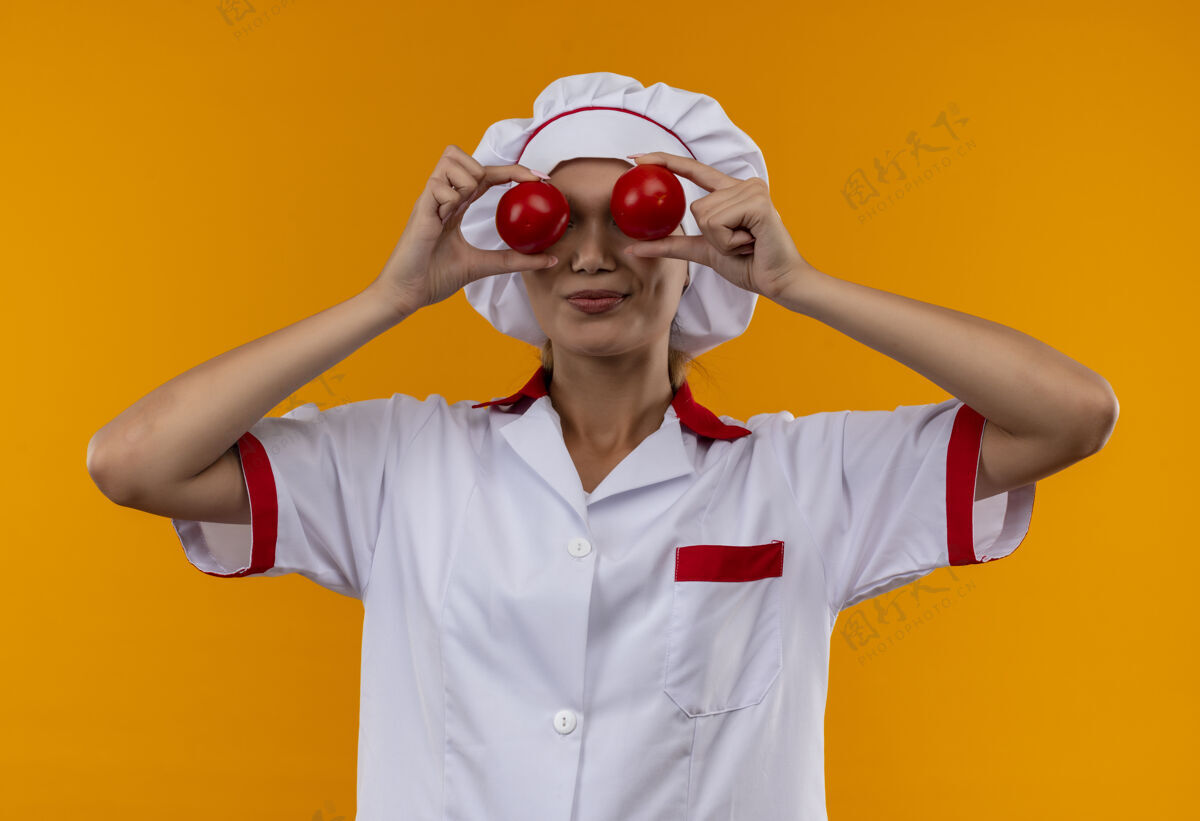 年轻高兴的年轻厨师穿着厨师制服的女厨师在孤立的橙色墙上涂满了西红柿的眼睛封面女穿着